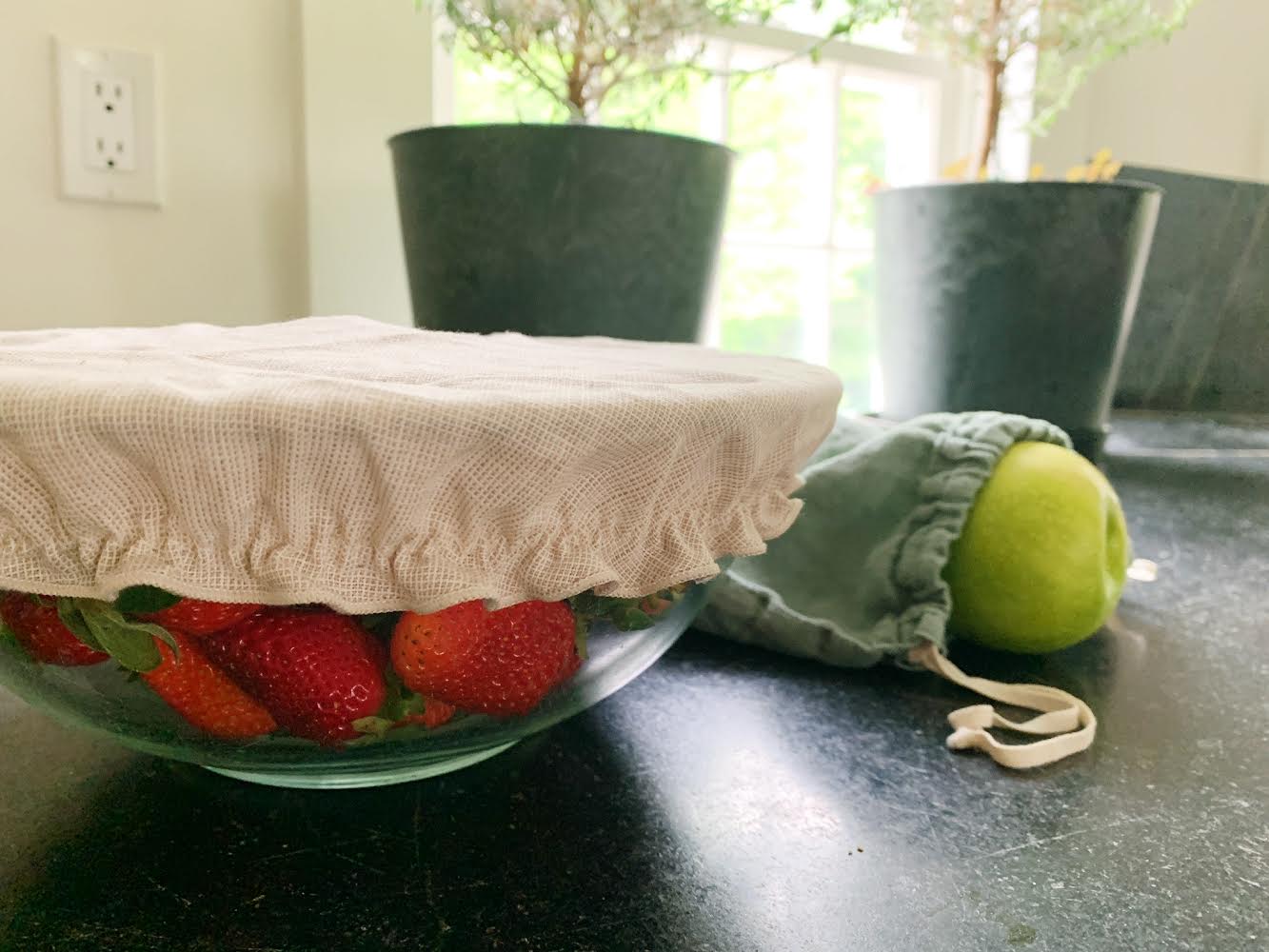What a reuse future includes. Reusable cloth bowl cover covering up of strawberries. Another reusable bag is in the background with an apple inside.