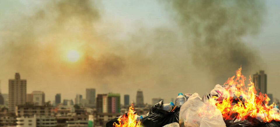 Pile of trash on fire and smoking in the foreground. Cityscape in the background.