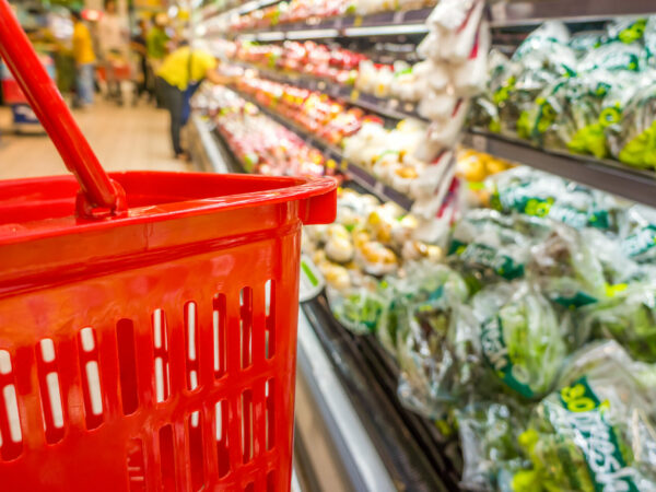 Close-up of red grocery basket next to an aisle of vegetables packed in plastic