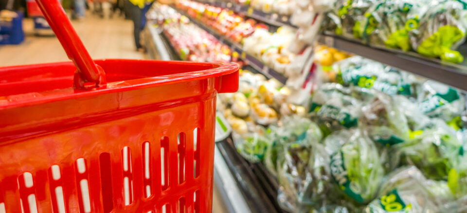 Close-up of red grocery basket next to an aisle of vegetables packed in plastic