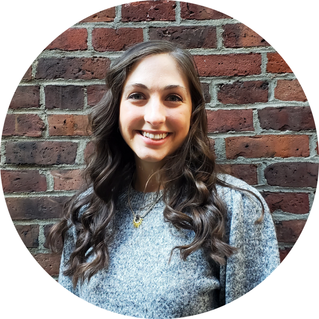 Headshot of a woman with long, dark brown hair standing in front of a brick wall. She's wearing a grey sweater and smiling.