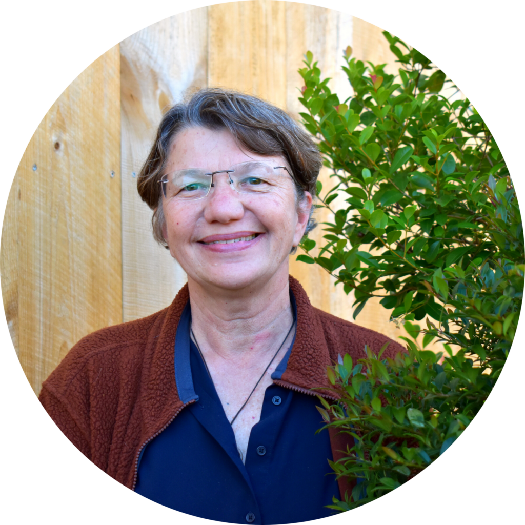 Headshot of a woman with short, brown hair standing in front of a barn door and next to a plant. Wearing a blue shirt, a rust colored sweater, and smiling.