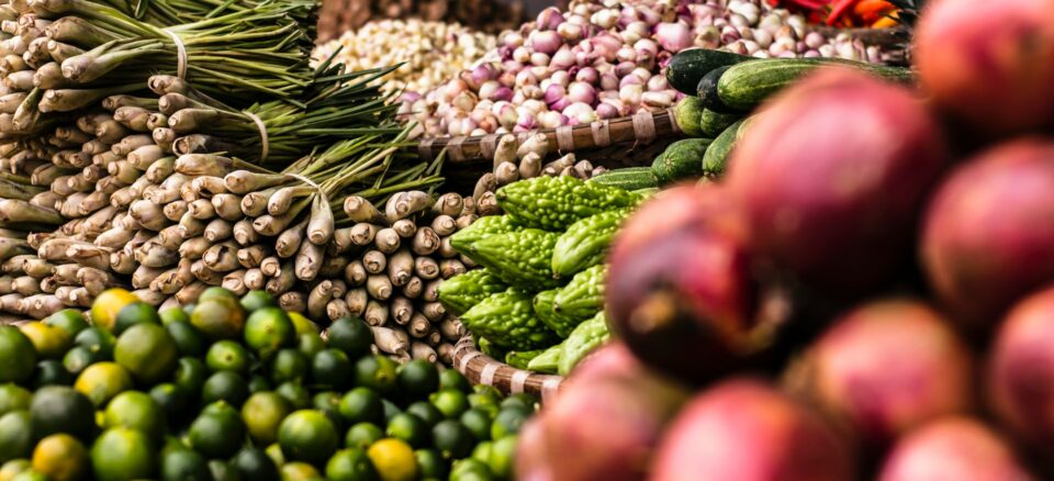 Market with piles of fresh produce including scallions, limes, shallots, and more.